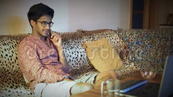 戴眼镜的印度男人用电话讨论商务问题坐在沙发前的笔记本电脑视频的预览图