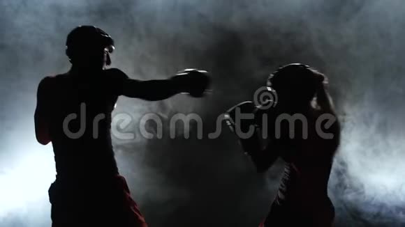 一个戴着头盔和拳击手套的女孩在黑暗中在拳击场上打的家伙剪影烟雾背景慢慢视频的预览图