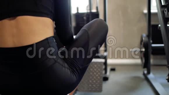 穿着黑色紧身裤的黑发少女坐在健身房的长凳上锻炼后休息视频的预览图