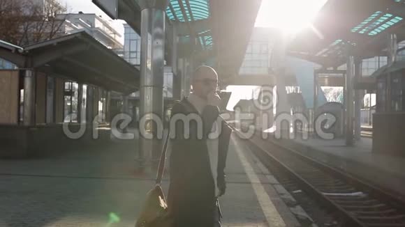 穿着雨衣和太阳镜的商人在火车站的站台上抽烟等火车视频的预览图