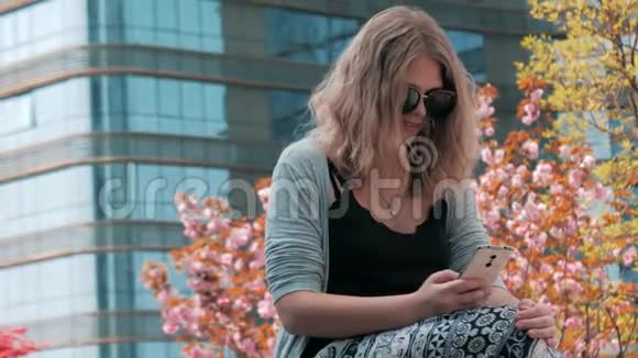 一位年轻漂亮的欧洲女孩戴着太阳镜坐在街上用智能手机连接摩天大楼视频的预览图