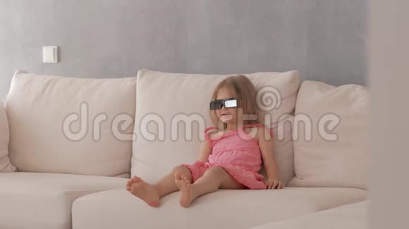 一个穿着粉红色裙子的小女孩坐在沙发上用3D眼镜专注而有感情地看电视视频的预览图