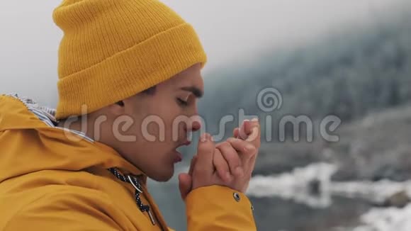 徒步旅行者手吹寒冷的天气站在美丽湖泊附近的雪山上的年轻旅行者视频的预览图