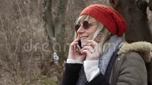 年轻漂亮的女人戴着红帽子坐在树旁用手机说话视频的预览图