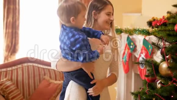 年轻母亲抱着她蹒跚学步的小儿子和在客厅装饰圣诞树的4K镜头视频的预览图