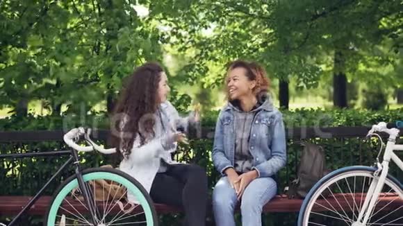 快乐的女孩坐在公园的长凳上聊天和笑自行车和背包是可见的现代生活方式城市视频的预览图