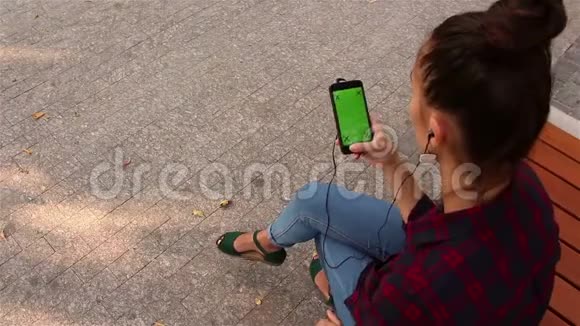 年轻漂亮的女孩坐在公园的长凳上在智能手机上浏览社交网络视频的预览图