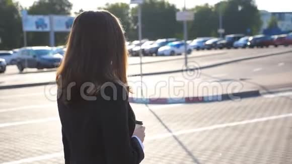 阳光明媚的一天年轻女商人带着一杯咖啡在汽车停车场附近散步女商人去上班视频的预览图