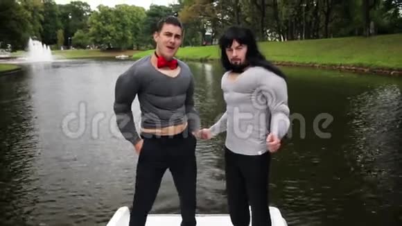 两个穿着假肌肉胸衣和双臂垫服装的迷人男子站在船上视频的预览图