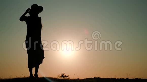 一个年轻女子戴着一顶帽子穿着一件长长的连衣裙在夕阳的映衬下在田野里捋头发视频的预览图