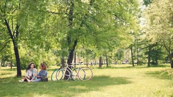 两个漂亮的女孩朋友正在公园的草坪上谈笑风生天气晴朗夏天现代自行车视频的预览图