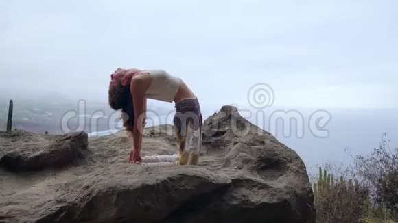 女孩在岩石上练习瑜伽对抗蓝天和蔚蓝的海洋女人站在石头上站在桥上的姿势视频的预览图