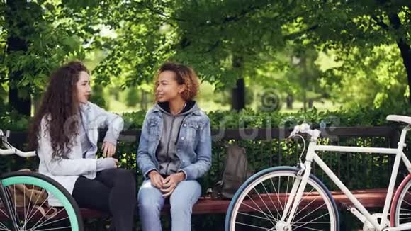 快乐的女性朋友正在谈论坐在镇上公园的长凳上看着自行车和袋子的新闻人民视频的预览图