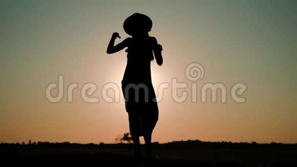 一个年轻女子戴着一顶帽子穿着一件长长的连衣裙在夕阳的映衬下在田野里捋头发视频的预览图