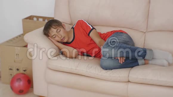 年轻人睡在沙发上穿着红色t恤和蓝色牛仔裤的白人少年在米色皮革沙发上睡觉视频的预览图
