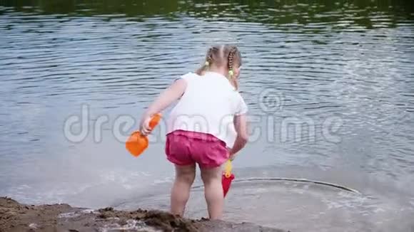 小女孩用水桶从河里舀水动作缓慢孩子们在沙滩上玩自然游戏视频的预览图