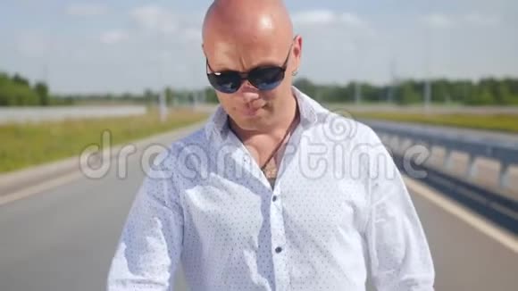 一个戴太阳镜的秃头男人在路上唱歌和散步的肖像中弹视频的预览图