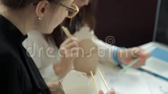 两个戴着眼镜的年轻女孩坐在笔记本电脑旁的咖啡馆里吃着中国面条讨论着这张照片视频的预览图