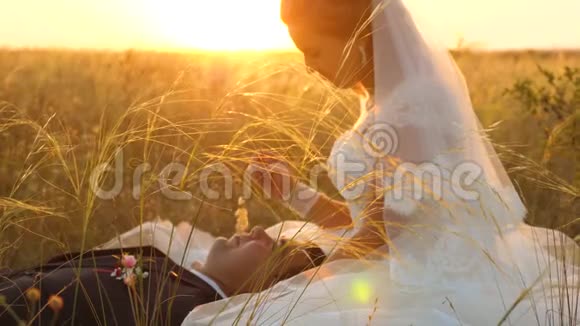 新娘当面用草柄抚摸新郎幸福的家庭躺在美丽的夕阳下的田野里年轻女子视频的预览图
