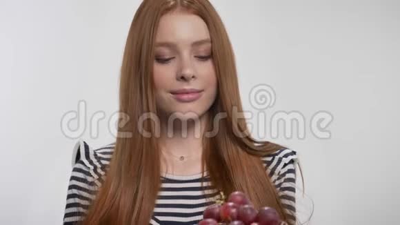 年轻可爱的生姜女孩拿着葡萄献祭看着镜头白色背景视频的预览图