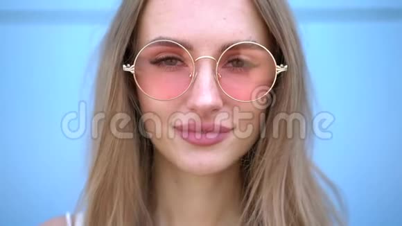 美丽时尚微笑女人的肖像有飞扬的发型蓝色背景上的粉红色霓虹太阳镜伤亡视频的预览图