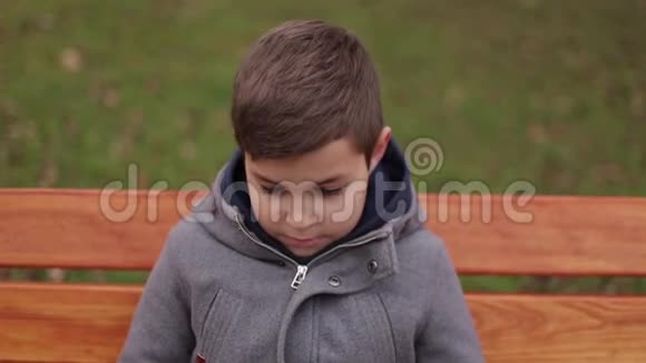 小男孩坐在公园的长凳上对着镜头微笑穿夹克的帅哥秋天慢动作视频的预览图