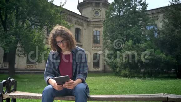 年轻的帅哥留着长长的卷发在平板电脑上打字在大学或校园附近的公园里坐在长凳上视频的预览图