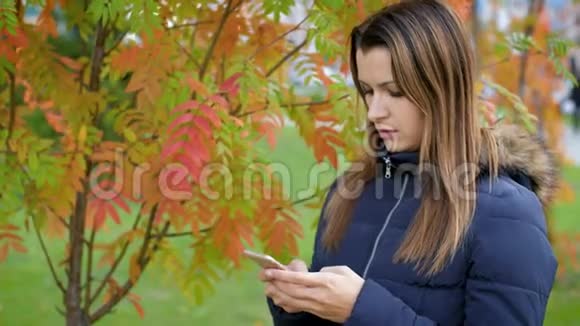 一个年轻的女人拿着一部手机站在鲁文脸红的灌木丛前女孩用智能手机站在视频的预览图