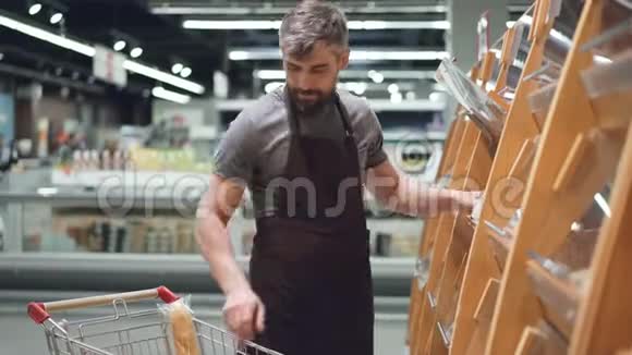 忙碌的推销员正把面包放在食品店的面包店的货架上留胡子的家伙穿着围裙出售视频的预览图
