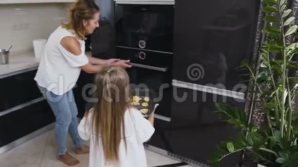 小女孩穿着白色连衣裙长发把烤盘和饼干放在烤箱里幸福的年轻家庭正在做饭烘焙视频的预览图