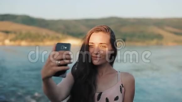 一个美丽的女孩用手机自拍站在河山前半身像视频的预览图
