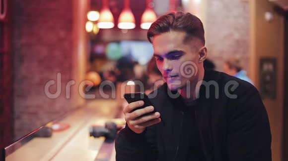 年轻的魅力男子与美丽的微笑是使用智能手机在晚上他坐在靠近霓虹灯的酒吧或餐馆里视频的预览图