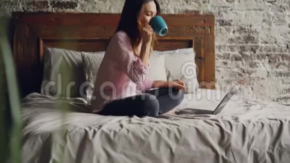 漂亮的年轻女性穿着休闲装坐在双人床上用笔记本电脑在Skype上和朋友聊天视频的预览图