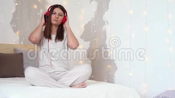 穿着睡衣听音乐的漂亮女孩戴着耳机坐在床上视频的预览图