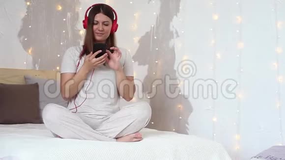 美丽的女孩听音乐耳机红色坐在床上放松使用手机总体计划视频的预览图