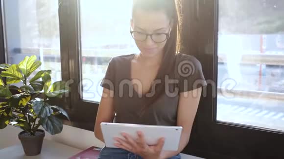 漂亮的年轻商务女性坐在办公室的窗户旁边使用她的数字平板电脑视频的预览图