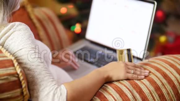4K镜头女性坐在温暖的圣诞树旁边的扶手椅上浏览圣诞礼物的互联网视频的预览图