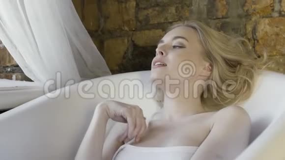 一幅美丽的金发女孩的肖像她穿着白色无肩带上衣坐在红砖墙背景上的空浴室里视频的预览图