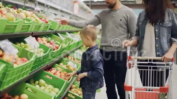 年轻的家庭妈妈爸爸和小男孩正在超市里挑选水果触摸和闻它们然后放在手推车里视频的预览图
