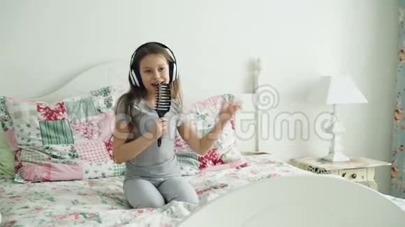 有趣可爱的小女孩戴着无线耳机跳舞梳着梳子在度假的早晨坐在床上玩得开心视频的预览图