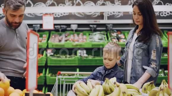 妈妈爸爸和可爱的男孩在食品店里挑选水果孩子在闻香蕉把它们送给他的父亲男人是视频的预览图