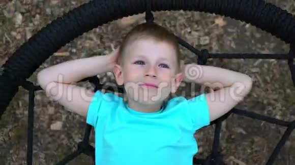 蓝眼睛的五岁男孩穿着蓝色t恤躺在一个圆秋千上双手仰起头抬头看着视频的预览图