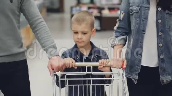 可爱的孩子在食品店里推着购物车他慈爱的父母在帮助他儿童青年家庭视频的预览图