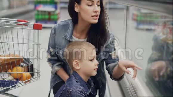 可爱的女人正在冰箱里为她可爱的小儿子选择冰淇淋用玻璃看食物指着物品视频的预览图