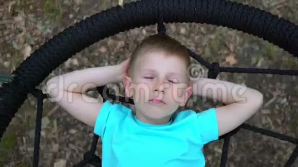 一个蓝眼睛的5岁男孩穿着一件蓝色T恤闭着眼睛做梦躺在一个圆圆的秋千上孩子微笑着视频的预览图
