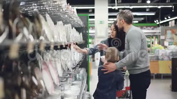 丈夫和妻子在超市的器皿部选择盘子他们的小儿子在帮助他们购买视频的预览图