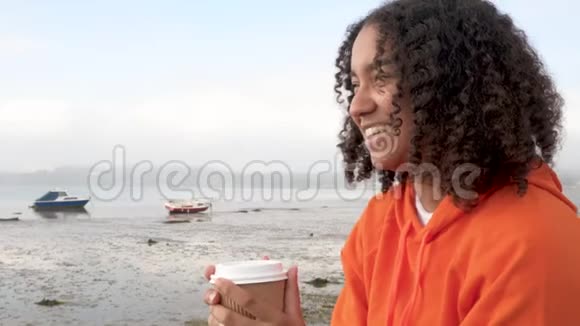 穿着橙色连帽衫在海边喝外卖咖啡的美国黑人少女少女视频的预览图