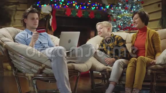 当圣诞礼物盒落在他的母亲和弟弟身上时男人在笔记本电脑上网购视频的预览图