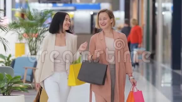 两个快乐的年轻女人慢吞吞地拎着销售袋穿过购物中心视频的预览图