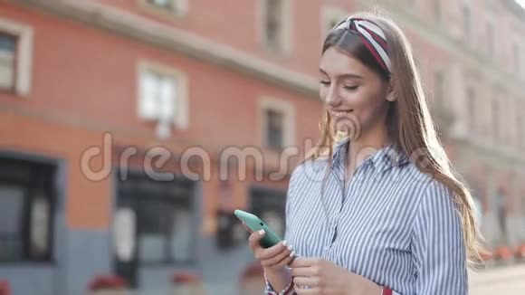 微笑的年轻女子穿着蓝色和白色条纹连衣裙用智能手机在老街上走来走去视频的预览图